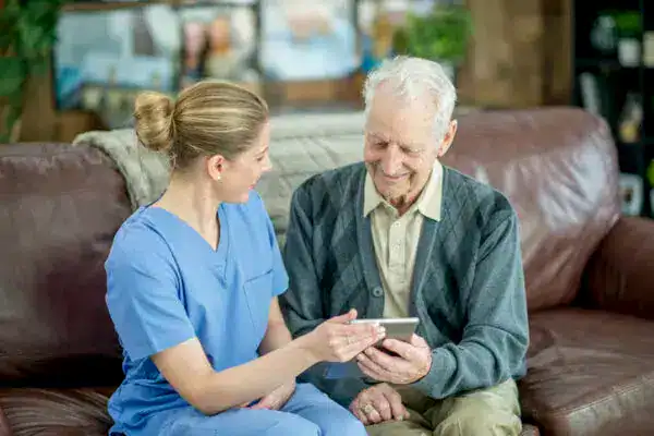 A nurse sits next to an elderly man on a couch, showing him something on a smartphone. Both are smiling, highlighting the warmth and connection of quality homecare services.