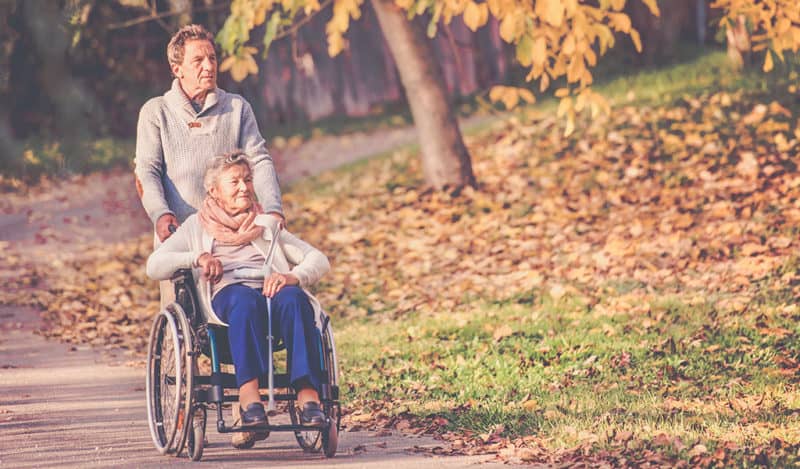 MAN PUSHING AN ELDERLY WOMAN IN A WHEELCHAIR DOWN A TREE-LINED PATH IN AUTUMN, PROVIDING ELDERLY HOME CARE.