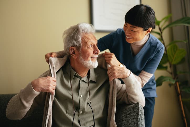 An elderly man, seated on a couch, is receiving companion care from a caregiver.