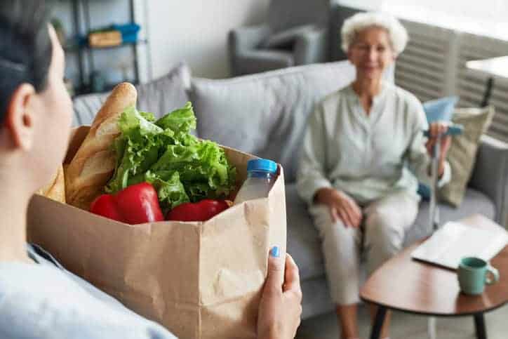 A person holding a bag of groceries, including bread, lettuce, and bell peppers, approaches an older woman sitting on a couch with a walking stick in her hand. This scene exemplifies the essence of errand running services offered through in-home care for the elderly.