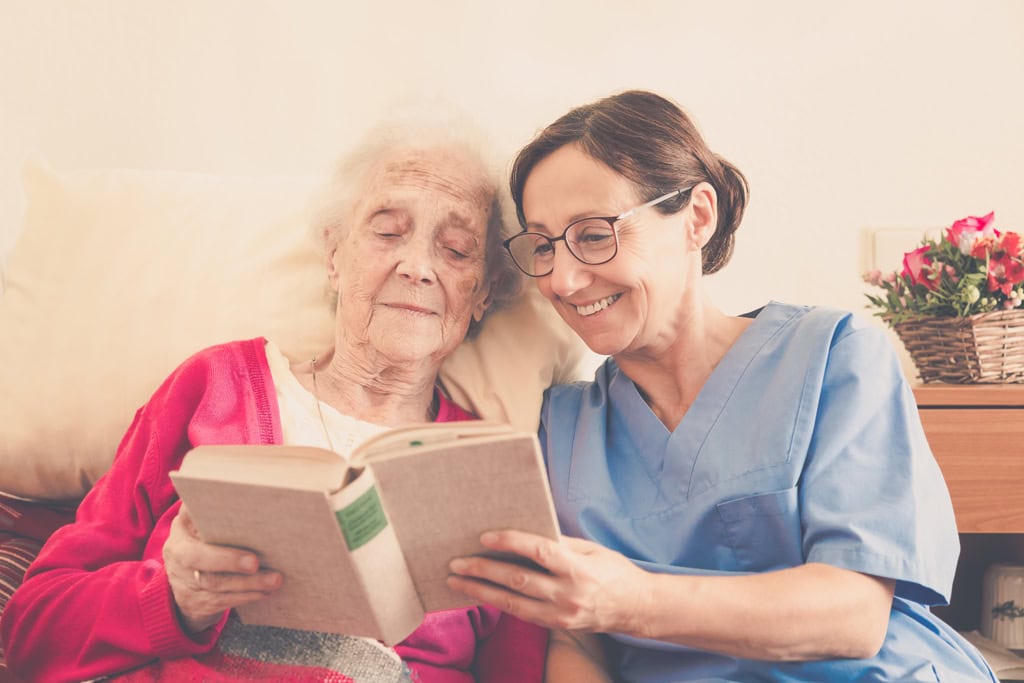 A caregiver attentively reads a book to an elderly woman in senior home care.