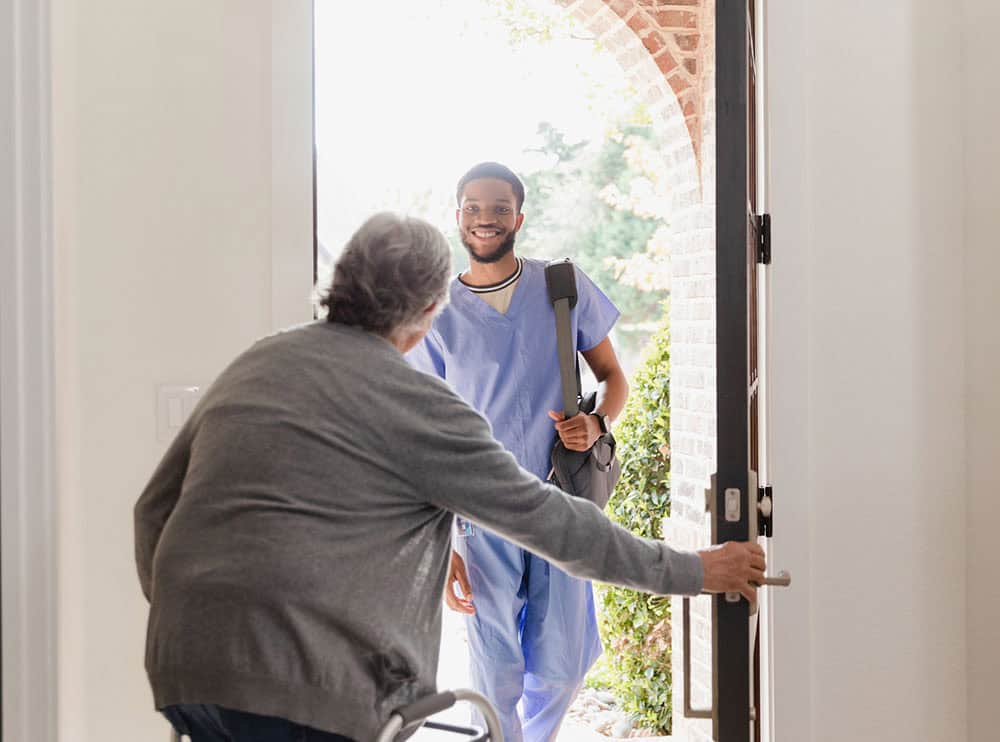 The senior adult man with a mobility walker opens the front door of his home for the young adult male physical therapist.