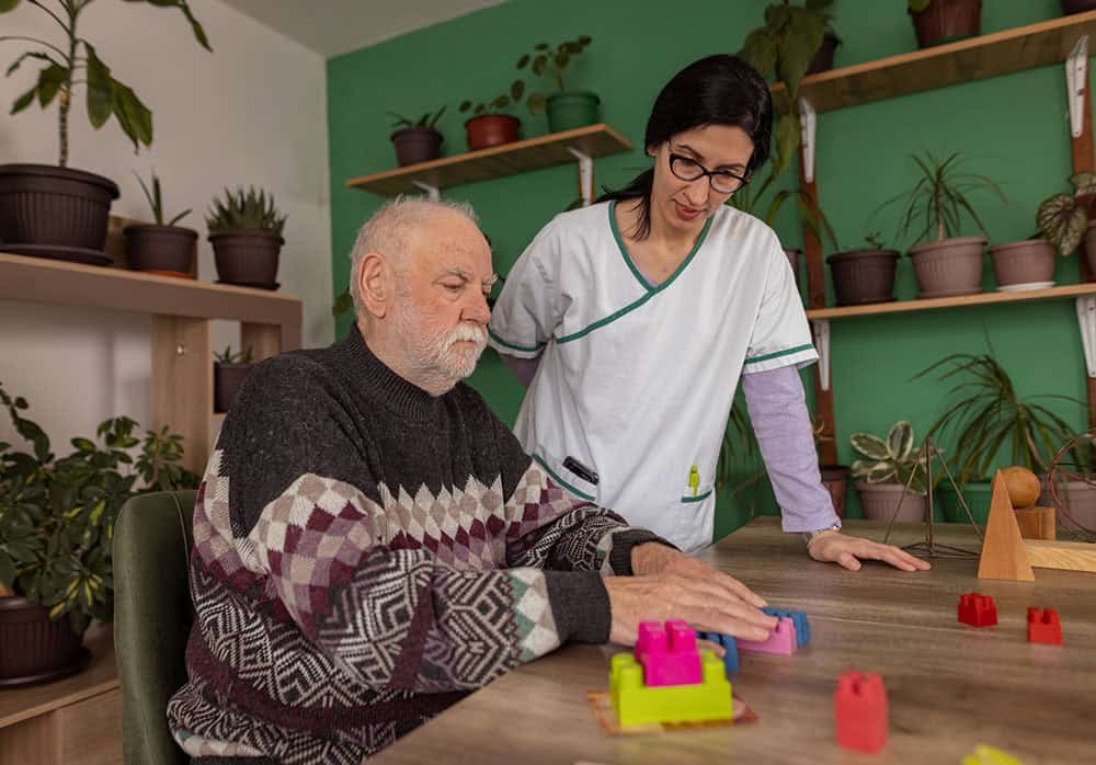 A senior man with dementia is playing with a colorful toy block while a female nurse is encouraging him during a toy therapy session.